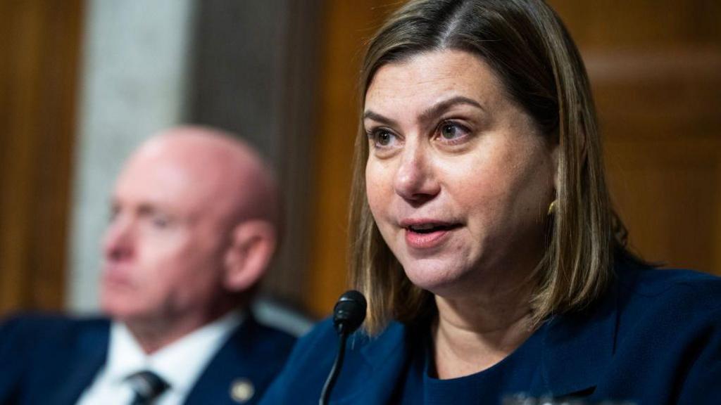 Michigan Senator Elissa Slotkin appears in Congress speaking into a microphone while Arizona Senator Mark Kelly sits behind her out of focus
