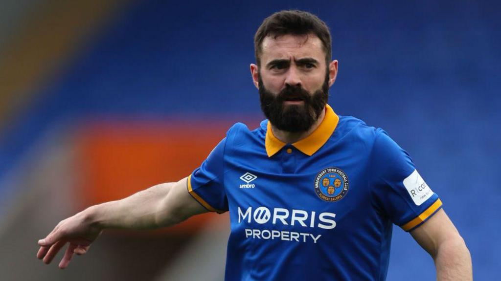 Jack Price points out an instruction to team-mates during a game for Shrewsbury Town. He is wearing a dark blue shirt with yellow edging on the sleeves.