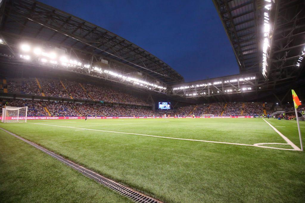 A view of a football stadium from the side of the pitch near the corner flag. There is a large stadium with lights around near the roof and a large screen in the far corner