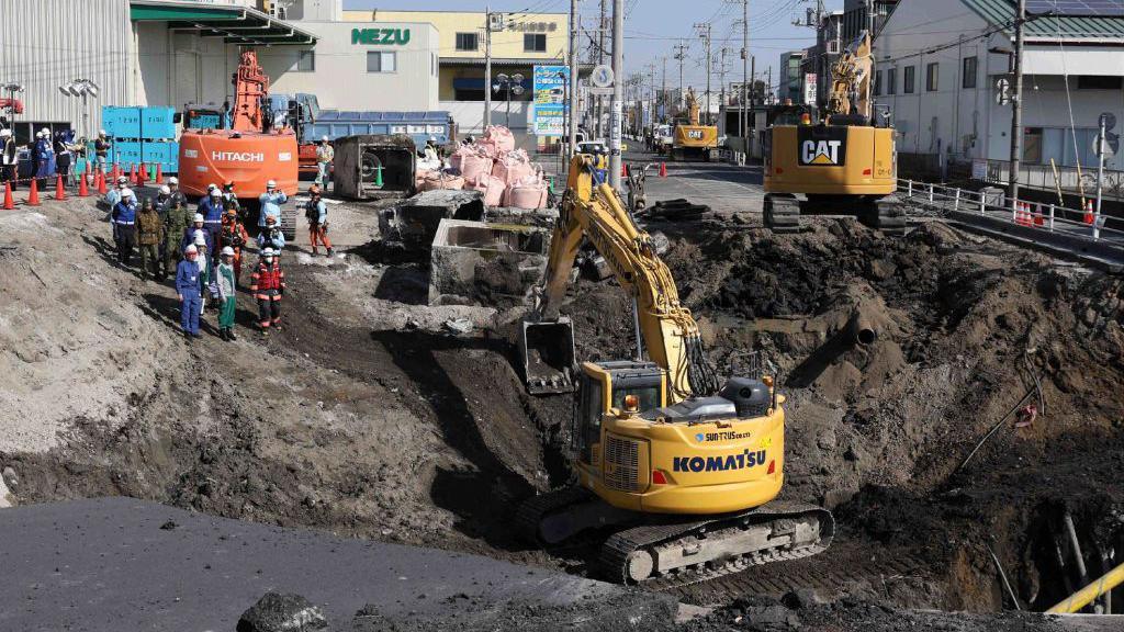 An excavator in a sinkhole, as a group of workers with helmets watch on