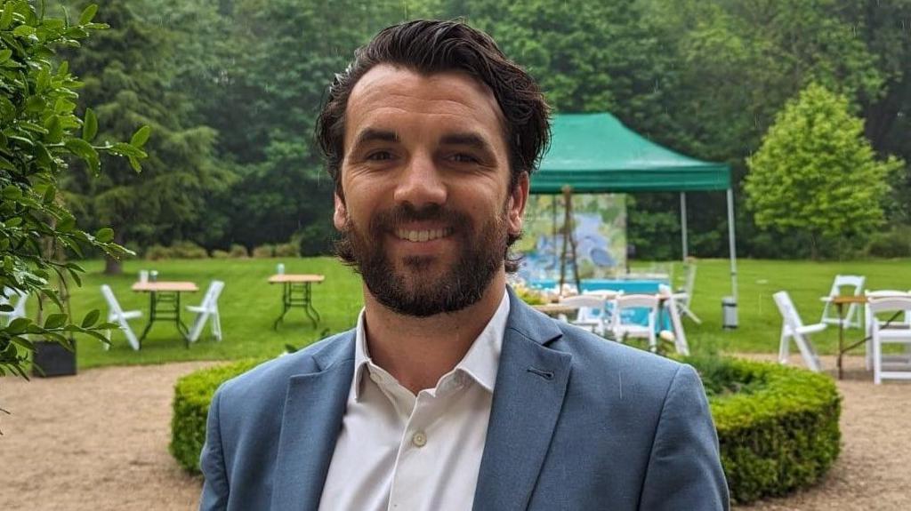 Man in a blue suit and white shirt smiling outdoors, with tables, chairs, and green trees in the background.