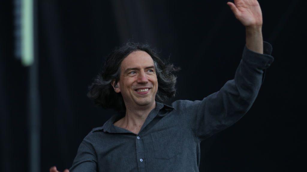 Gary Lightbody smiling and waving. He is wearing a grey shirt