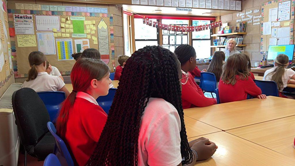 Children in a class wearing white tops or red jumpers are sat on blue plastic chairs. They are pictured from the back and are facing the front of the class where a woman is talking to them and giving a presentation using a computer.