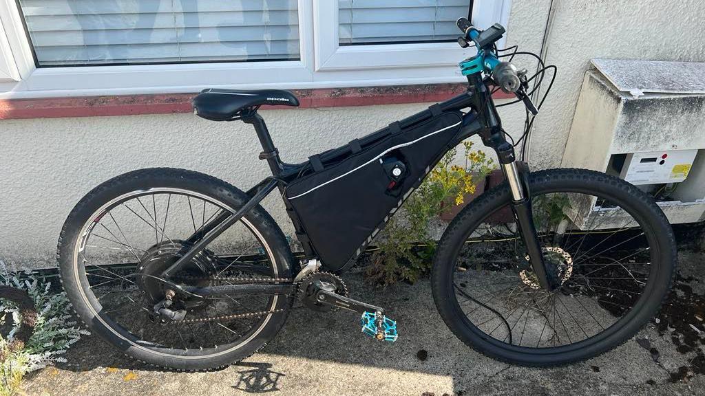 A black e-bike leant up against a windowsill outside a home, with blue handlebars and pedals.