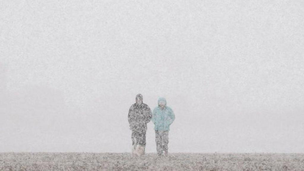 Two people in thick coats walk through falling snow across a flat landscape