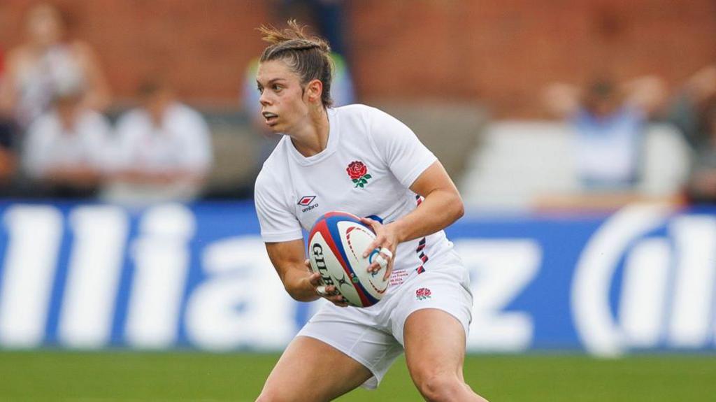 Helena Rowland in an all white England Rugby kit holding a rugby ball.