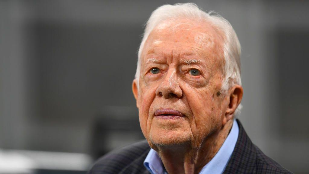 Jimmy Carter, an elderly, wrinkled man with white hair. He is wearing a blue-collared shirt with a black, checkered sports jacket