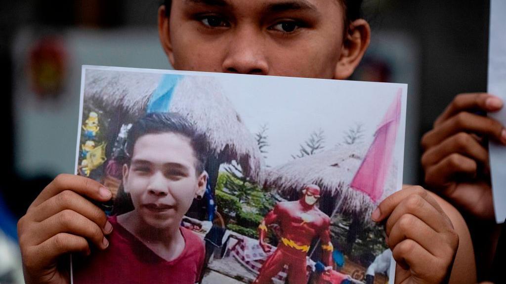 A supporter of Kian Delos Santos attends a protest outside the police station where three policemen involved in Kian's killing were assigned in Manila on November 29, 2018. He is holding up a photo of a smiling Kian in a red shirt, standing near a superhero figurine.