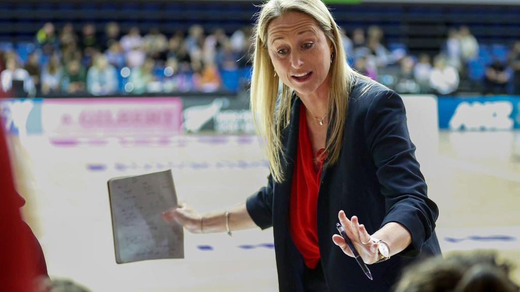 Jess Thirlby, as England Netball head coach, talking to her players during a break with a pen in her left hand and a piece of paper with notes on in her right hand.