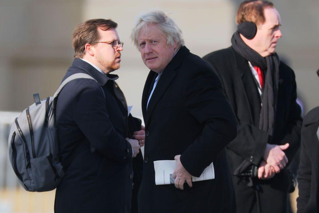 Boris Johnson standing outside in a coat, with two other men near him. He is looking across towards the camera and holding a piece of paper against his left side 