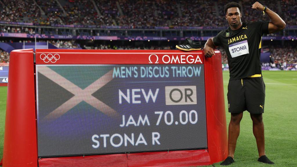 Roje Stona stands next to the board showing his Olympic discus record