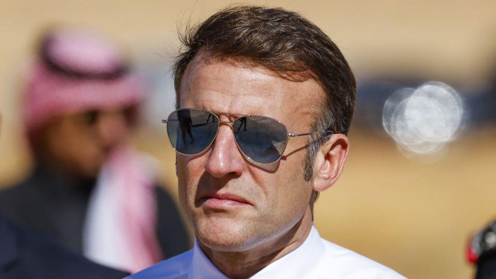 Emmanuel Macron during a visit to Saudi Arabia, wearing aviation sunglasses and a white shirt with a bit of a smirk on his face 