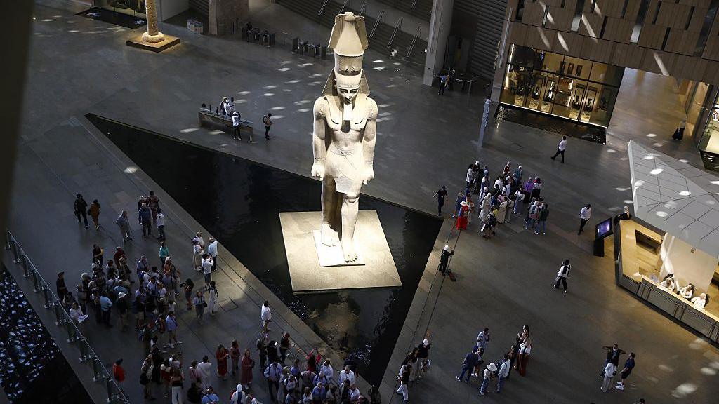View of inside the museum from above. A huge statue is seen in the middle of a foyer with lots of people walking past and some are seen sitting on benches. 