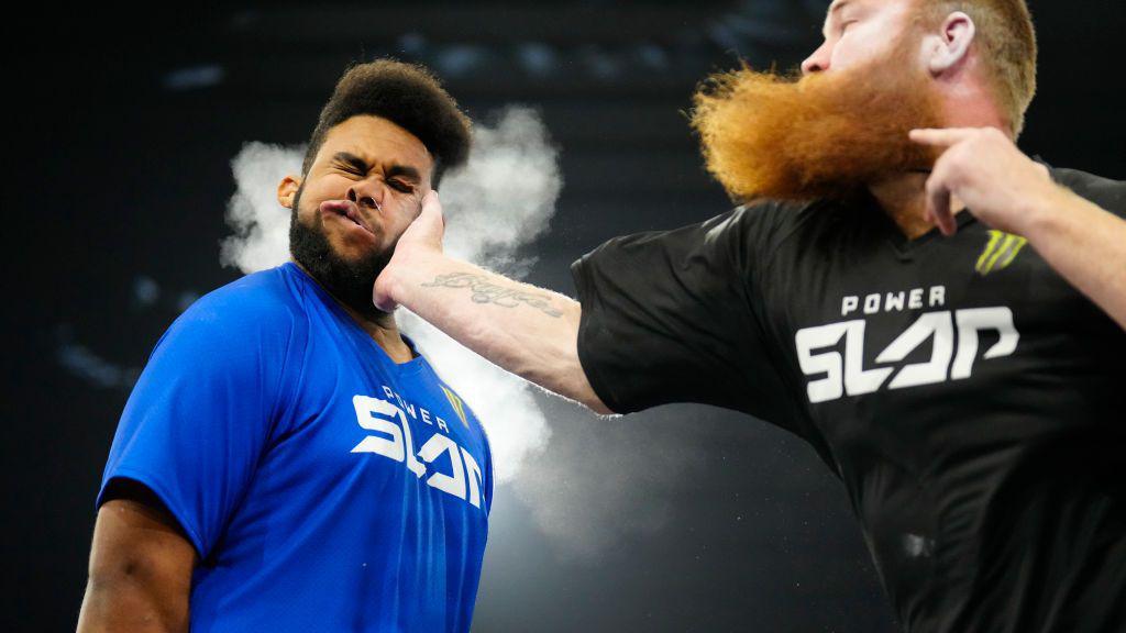 A man with a long ginger beard has his arm outstretched and he is slapping another man around the face in a competition. He is wearing a black T-shirt that says "power slap" on it. the other man's face is contorted due to being mid-slap, and he has brown hair and facial hair. He is wearing a blue top that reads "power slap"