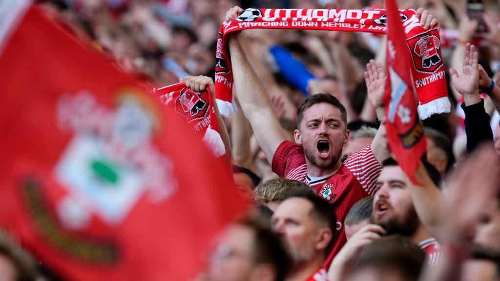 Southampton fans cheer their team on in the stands at Wembley in 2026. In the foreground is an out-of-focus flag, red in colour, with a Southampton FC badge on. In focus fans in Saints shirts cheer and hold aloft red scarves.