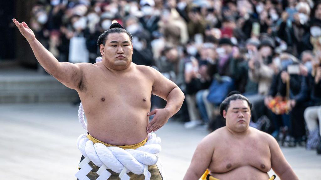 Hoshoryu, topless and in ceremonial dress from the waist down, with his arm raised. A crowd is in the background 