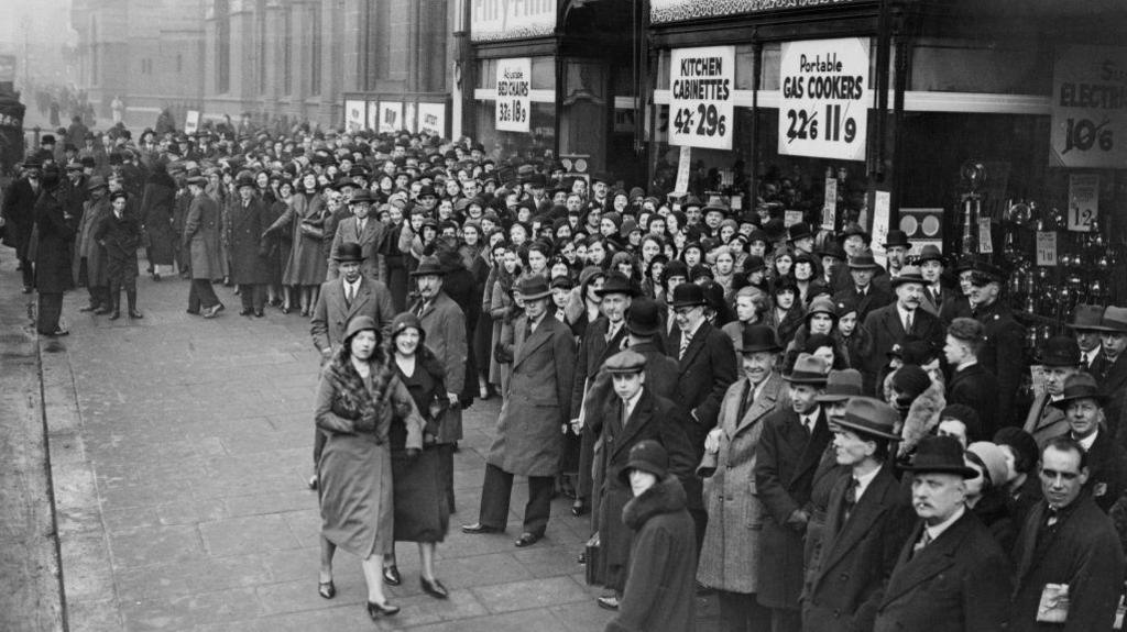 Crowds on the pavement outside Gamages department store in Holborn, London in February 1933. Signs on the storefront advertise offers for 'Adjustable Bed Chairs', 'Kitchen Cabinettes,' and 'Portable Gas Cookers.'  