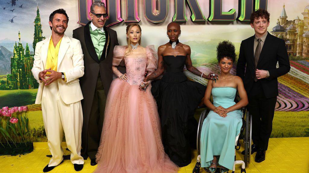 The image shows the principle case of Wicked. From left to right we see Johnathan Bailey who plays Prince Fiyero, Jeff Goldblum is the Wonderful Wizard of Oz, Glinda herself as portrayed by Ariana Grande and Elphaba is played by Cynthia Erivo. Marissa Bode plays Elphaba's sister Nessarose and Ethan Slater plays a munchkin called Boq. 