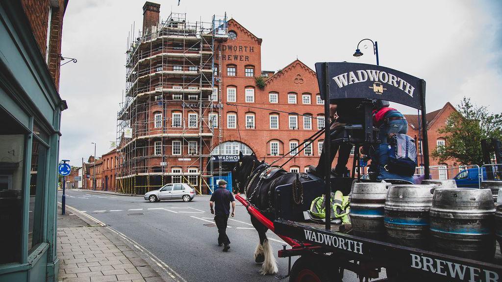 Side-view of Wadworth Cart & shire horse with barrels on the back, walking towards a large, Victorian red-brick building with Wadworth Brewery on the side.