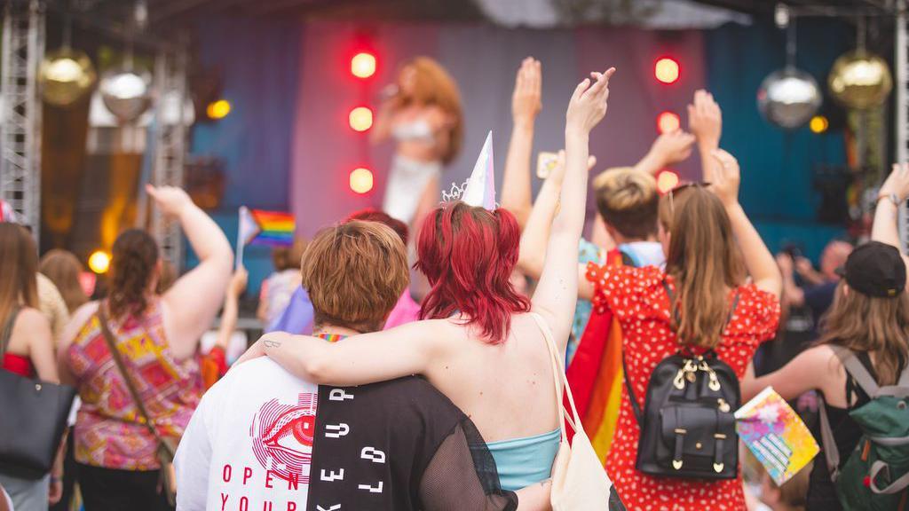 A crowd of people stood in front of a stage. Some people are holding rainbow flags. Others are holding their hands up in the air.