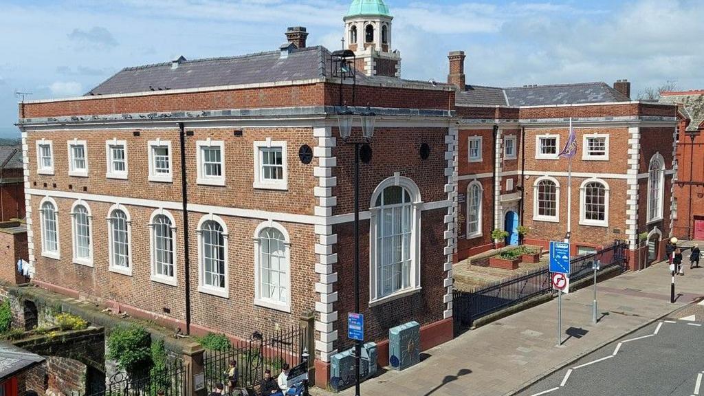 A large school building with two wings and a steeple with a turquoise domed roof and a black weathervane