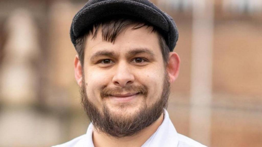 Image of a man with a beard smiling at the camera.  He has a grey flat cap on his head and is wearing a white shirt.