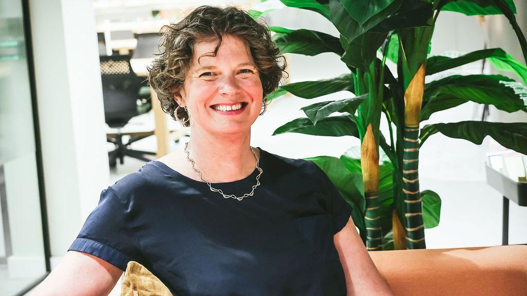 Kate sitting in an armchair with short hair smiling at the camera. There is a plant behind her and what looks like office space.