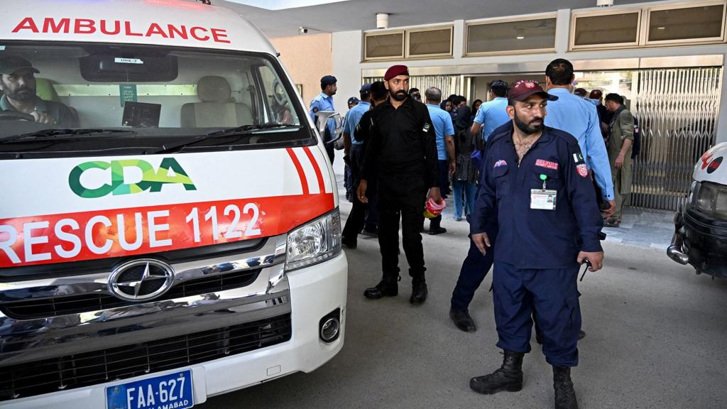 An ambulance is parked outside the hospital next to security staff and onlookers