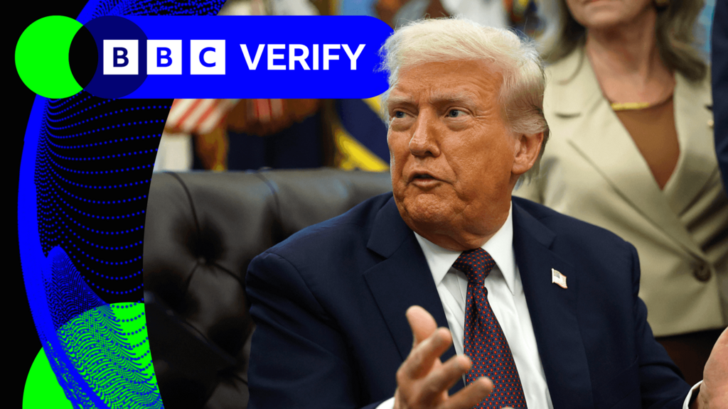 US President Donald Trump speaks to reporters in the Oval Office. He is wearing a dark suit with an American flag pin. Staffers can be seen standing in the background of the shot.