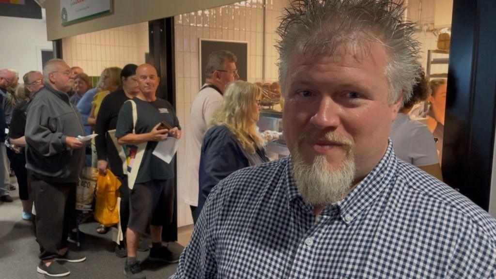 A man with a beard, wearing a blue and white checked shirt, is standing in front of a queue in a market.