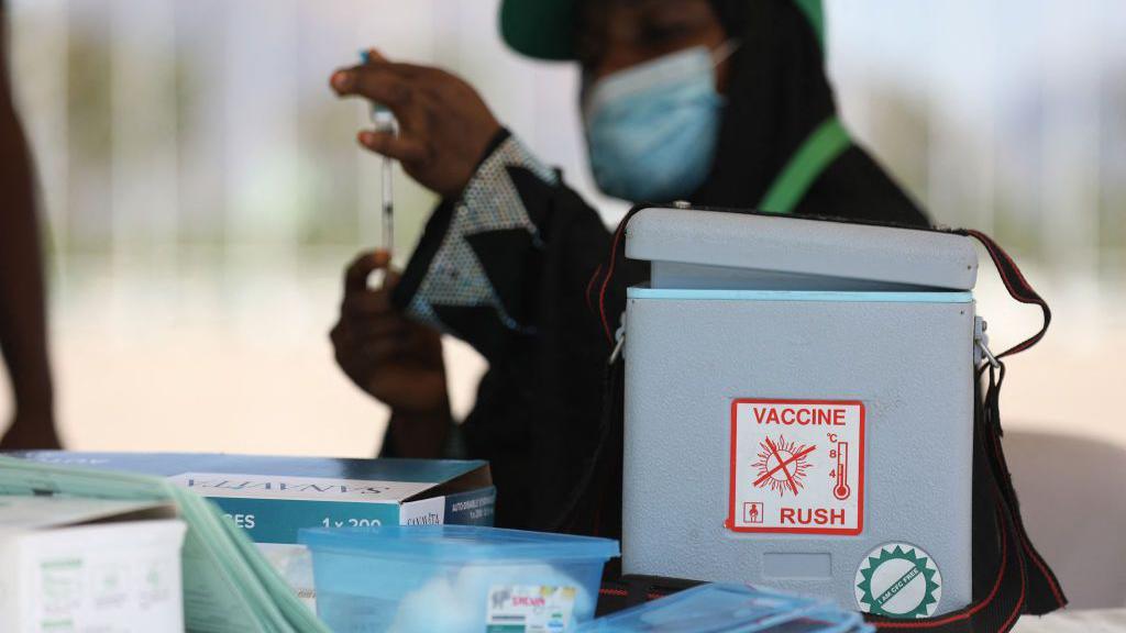 This image shows a vaccine storage and preparation area, with a portable insulated cooler prominently positioned in the foreground. A label on the cooler reads “VACCINE” and includes a temperature graphic, indicating the importance of maintaining a controlled cold chain. Nearby are medical supplies, including boxes, trays, and packaged equipment arranged on a work surface. In the background, a person wearing a face mask is holding a syringe upright, appearing to prepare or inspect it before use. The shallow depth of field keeps the cooler and vaccine-related equipment in sharper focus while the background remains blurred. The overall scene emphasizes the handling, storage, and preparation of vaccines in a healthcare or immunization setting.