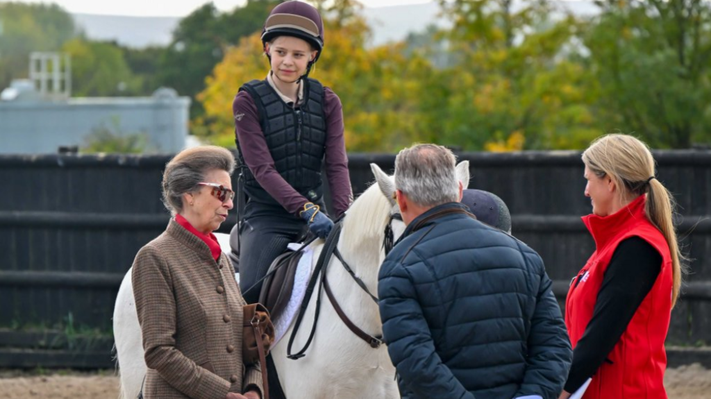 Princess Anne standing beside a white horse with a teenage girl sitting on the horse, talking to a man and woman with their backs to the camera