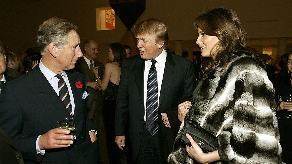 A younger King Charles, wearing a red poppy lapel pin and holding a drink, chats to Donald Trump and Melania. Drump wears a dark suit, and Melania is wearing a big black-and-white fur coat and carrying a black Chanel clutch