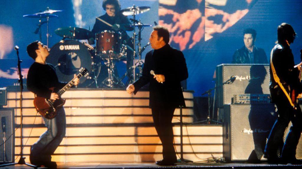 Tom JONES on stage with STEREOPHONICS, Kelly and Sir Tom are up front together look towards each other as they sing and Kelly plays guitar. You can also see the drummer keyboardist and the bas player is looking away.