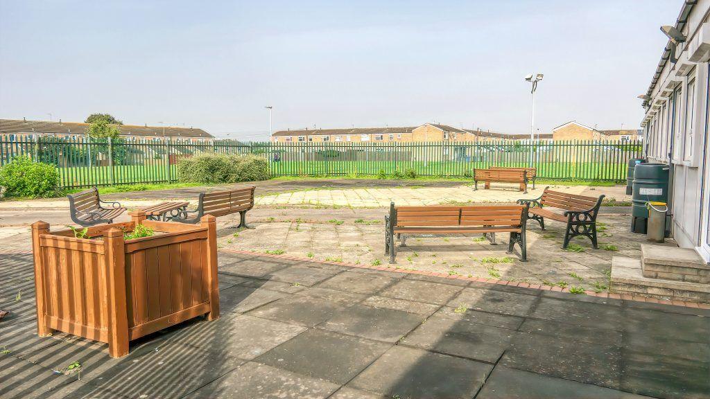 A large concrete paved area is fenced in. Benches are dotted around the area and a single planter sits in the foreground.