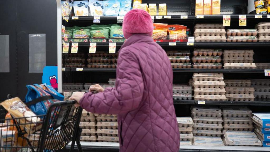 A woman in a purple puffy coat pushes a shopping cart past eggs in the grocery store