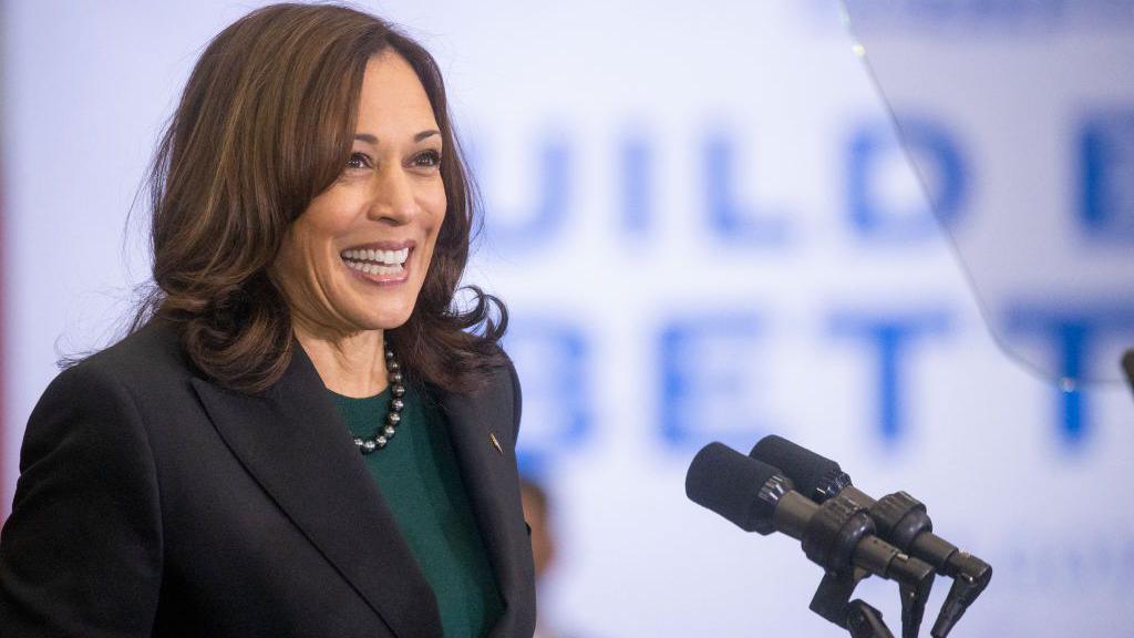 Kamala Harris is grinning widely while speaking into two microphones. Her brown hair is curled and she is wearing a black blazer, green top and a beaded necklace. In the photo she is framed to the left with a blurred blue and white sign in the background.