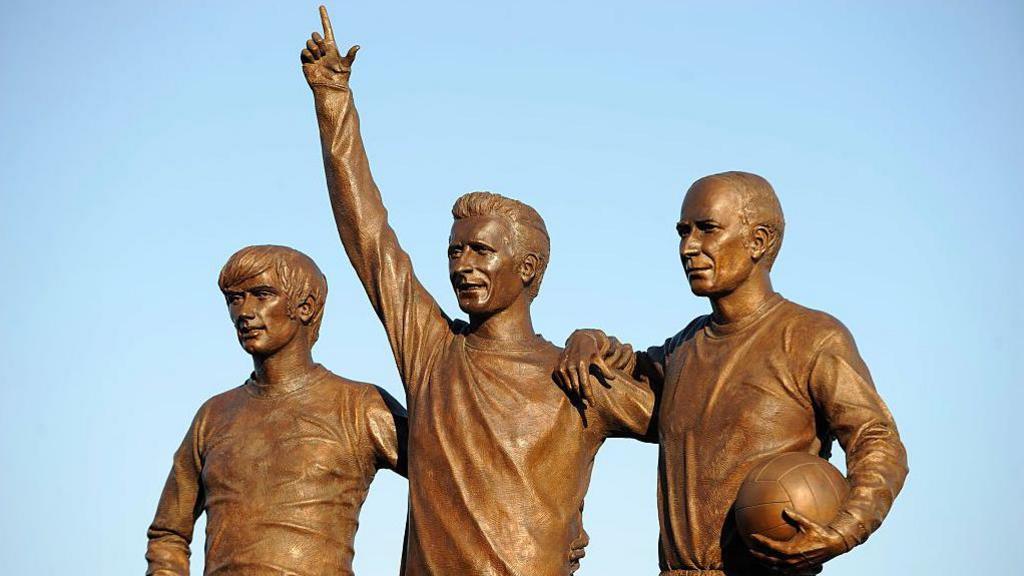 A bronze statue of three men in football strips. The one in the middle raises his hand to point at the sky and the man on the far right holds a football.