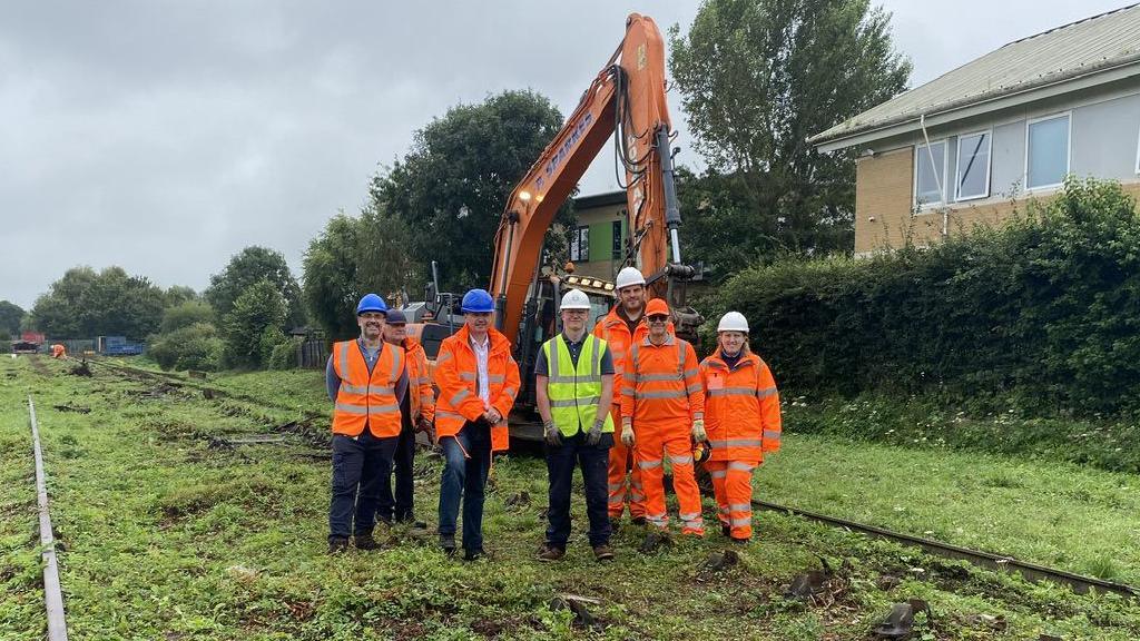 Several people in high-vis overalls standing in front of a digger