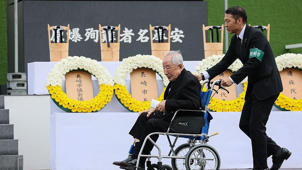Bomb survivor, Hiroshi Nishioka, who is very elderly and in a wheelchair, is pushed by another man, while wreaths of flowers are in the background.