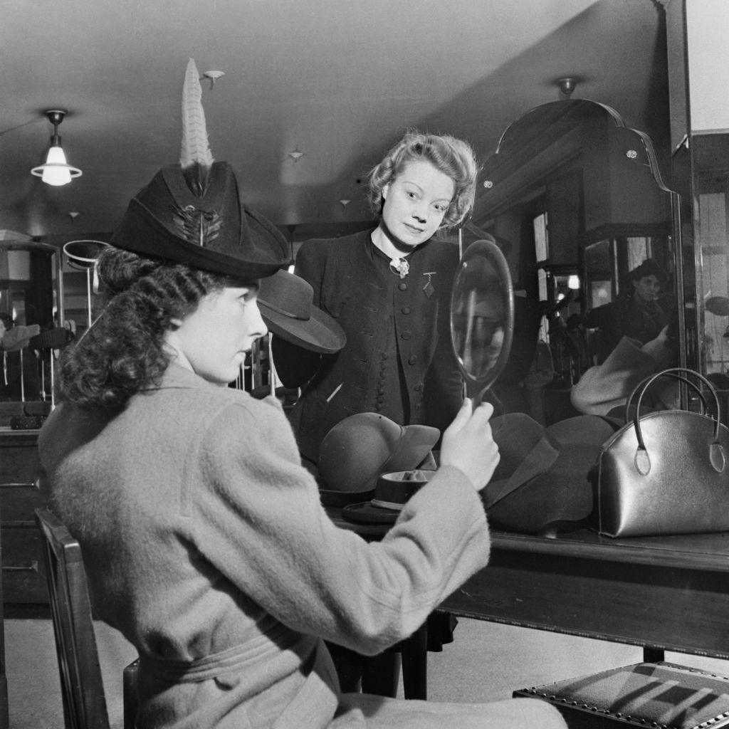 London Carries On: Shopping In Wartime London, 1942, A female customer tries on a new hat in the millinery department of Bourne and Hollingsworth on London's Oxford Street. She is examining her appearance in a mirror as the sales assistant looks on, circa 1942. (Photo by Ministry of Information Photo Division Photographer/ Imperial War Museums via Getty Images)