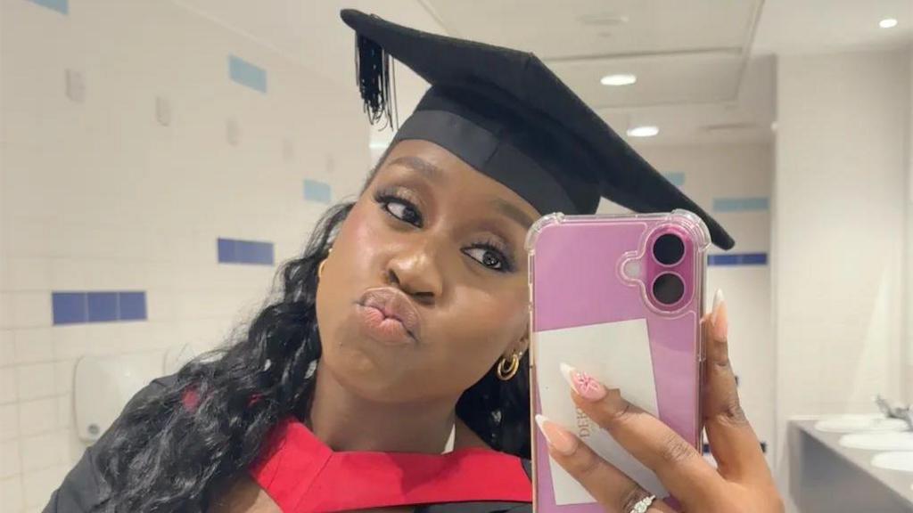 A young woman taking a mirror selfie with a pink phone, in her graduation cap and robes