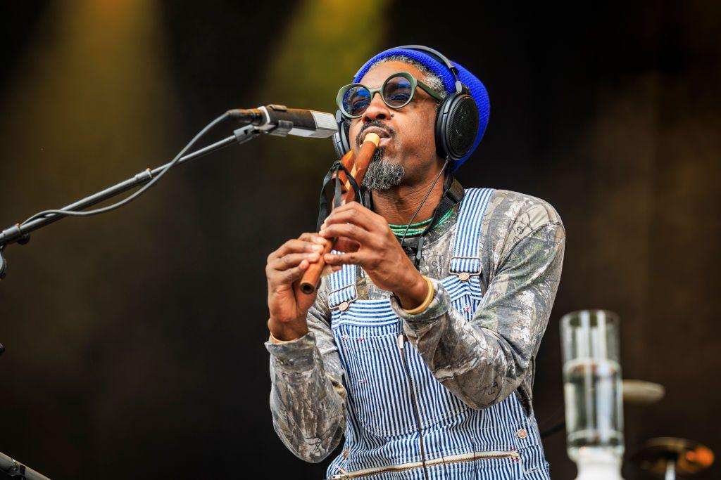 André 3000 plays a wooden wind instrument on stage in dark glasses and striped dungarees