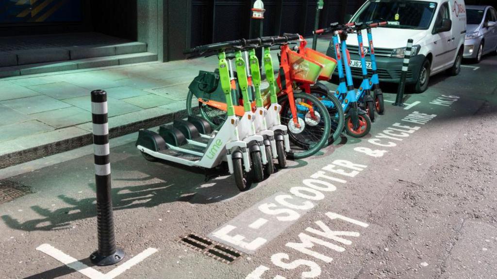 A row of rental e-scooters and bikes from Lime, Dott, and other providers is parked in a designated bay marked “E-scooter & cycle hire only” on a city street next to a white van.