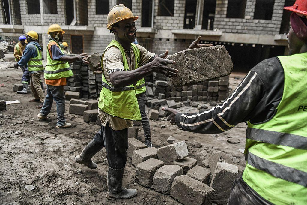 Men smile as they pass large bricks along to their colleagues on the building site. Behind them are building in their early stages.