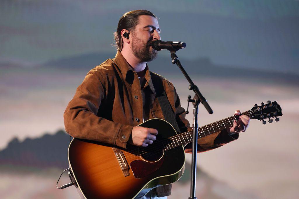 Noah Kahan in a brown suede jacket singing into a microphone holding a black and brown acoustic guitar. 