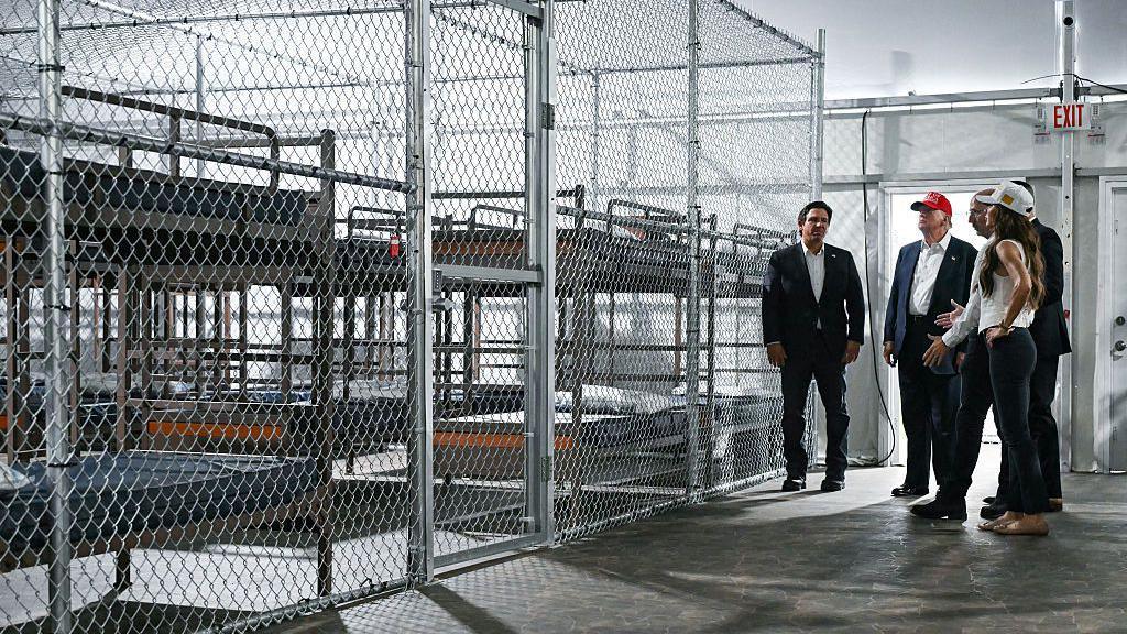 DeSantis, Trump in red cap, and Noem in white T-shirt and jeans in warehouse-like building next to rows of bunk beds surrounded by chain-link fencing