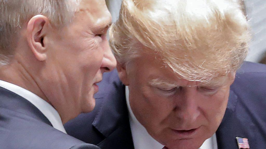 Vladimir Putin appears to speak directly into the ear of Donald Trump in a close-up from a summit in Vietnam in November 2017. Trump, wearing a US flag pin on his lapel, is looking down and is seen from the front, while Putin is in profile. 
