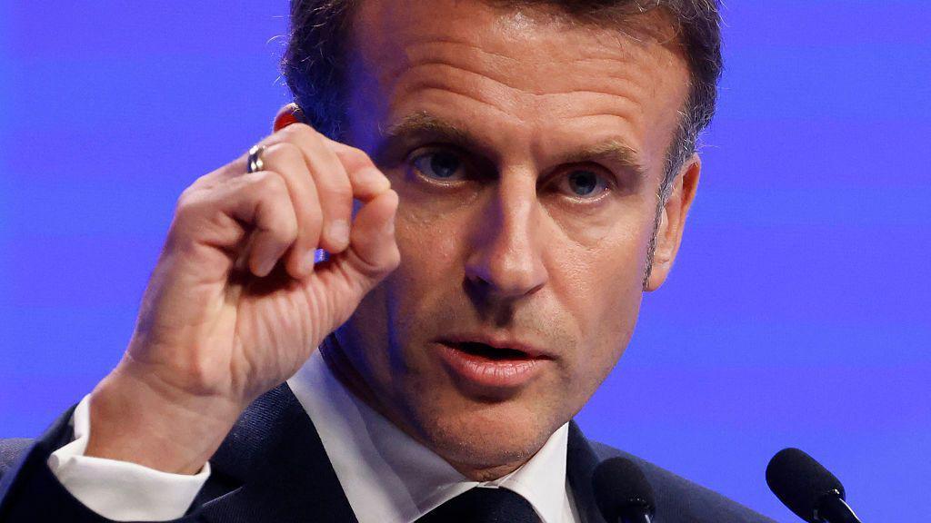 Emmanuel Macron gestures as he speaks at a podium in front of a blue screen.