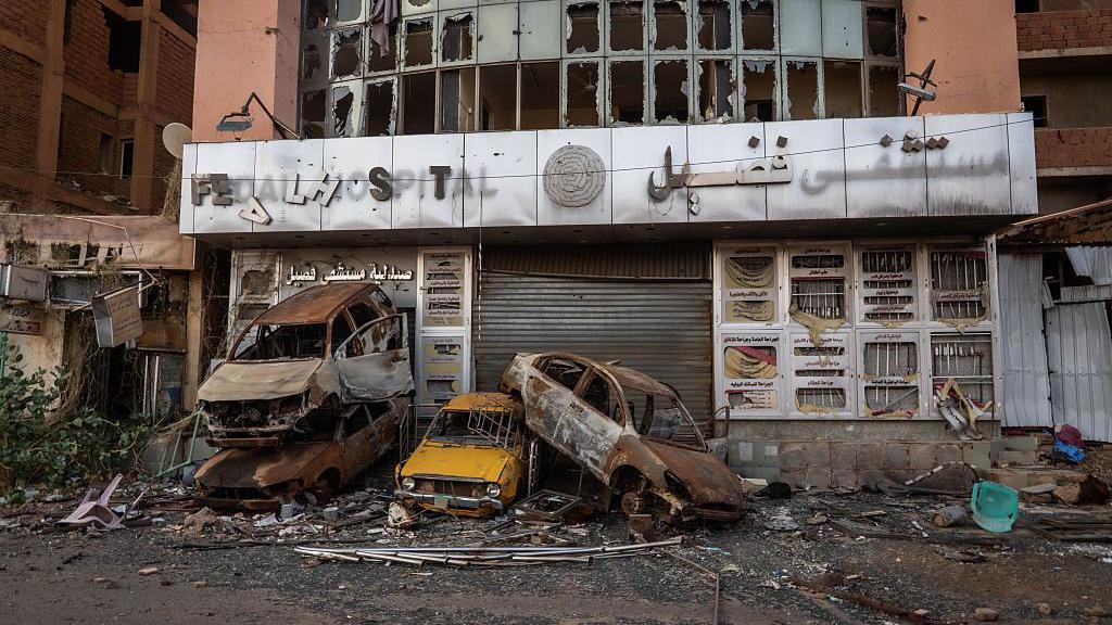 Burnt-out vehicles are piled in front of derelict building destroyed by conflict. The windows are blown out and a broken sign says 'hospital' in English.